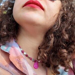 Side angle outdoors: pink beads and floral-stamped diamond spacers ring the base of the neck, the teardrop tipped forward against the skin, garden greenery behind.