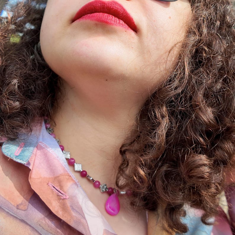 Side angle outdoors: pink beads and floral-stamped diamond spacers ring the base of the neck, the teardrop tipped forward against the skin, garden greenery behind.