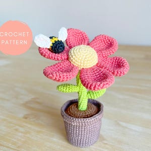 Może przedstawiać: Ręcznie robiony kwiat na szydełku z różowymi płatkami, żółtym środkiem i zieloną łodygą, w szarej doniczce. Mała pszczoła na szydełku spoczywa na kwiatku. Obraz zawiera tekst "CROCHET PATTERN" w różowym okręgu.