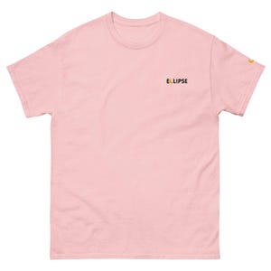 Puede incluir: Una camiseta rosa claro con la palabra "ECLIPSE" bordada en negro, amarillo y morado en el pecho. Un pequeño diseño amarillo está en la manga izquierda. La camiseta está hecha de un material suave.