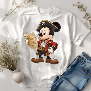 May include: White t-shirt featuring a cartoon pirate Mickey Mouse holding a treasure map. Mickey is dressed in a red coat, black pirate hat with a skull and crossbones, and brown boots. Blue jeans are visible in the background.