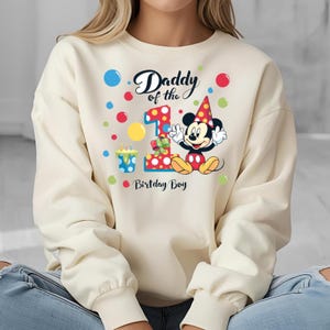 May include: Cream-colored sweatshirt featuring a colorful graphic of Mickey Mouse celebrating a first birthday. The design includes the text "Daddy of the 1st Birthday Boy" with colorful polka dots and a birthday cake.