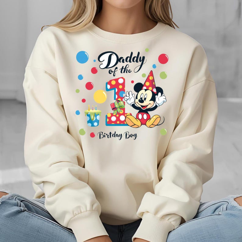 May include: Cream-colored sweatshirt featuring a colorful graphic of Mickey Mouse celebrating a first birthday. The design includes the text "Daddy of the 1st Birthday Boy" with colorful polka dots and a birthday cake.