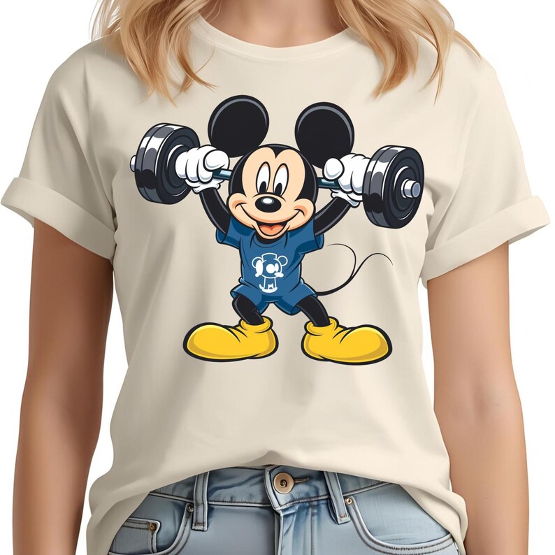 May include: A cream-colored t-shirt featuring a cartoon Mickey Mouse character lifting weights. Mickey is wearing a blue outfit and yellow shoes. The weights are black and gray.