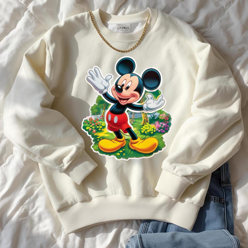 May include: Cream-colored sweatshirt featuring a colorful graphic of Mickey Mouse waving. The cartoon character is depicted in his classic outfit with red shorts and yellow shoes. A gold chain necklace is draped on the sweatshirt.