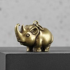 Puede incluir: Pequeña figura de elefante de latón detallada con un lazo para llavero. El elefante está sobre una superficie oscura, con un fondo gris texturizado. La figura tiene una superficie texturizada y es de color dorado antiguo.
