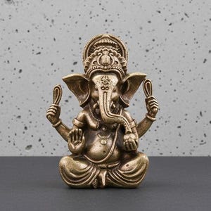May include: A small, bronze-coloured statue of the Hindu deity Ganesha. The statue shows Ganesha seated in a meditative pose, with multiple arms and an elephant head. The background is a speckled grey surface.