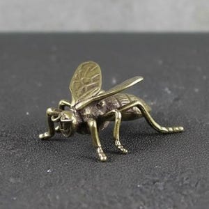 Puede incluir: Pequeña figura de abeja de latón. La abeja presenta detalles intrincados en sus alas y cuerpo, con seis patas y antenas. El latón tiene un aspecto ligeramente envejecido. La abeja está representada en posición de arrastre.