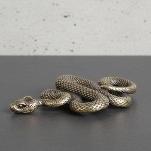 Könnte beinhalten: Detaillierte, metallische Schlangenskulptur mit strukturierter Oberfläche und detailliertem Kopf. Die Schlange ist aufgerollt und liegt auf einer dunklen Oberfläche, mit gesprenkeltem, grauem Hintergrund.