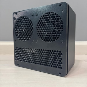 May include: A black, rectangular electronic device with two circular fan openings and a honeycomb-patterned vent. The device is mounted on a light-colored wooden surface, with a gray wall in the background.