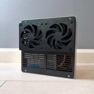 May include: A rectangular computer case component featuring a black 3D-printed mounting bracket with two integrated cooling fans. The lower section includes a black slatted grille revealing internal hardware, including a hard drive and colored cables.