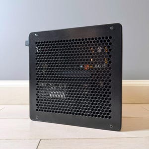May include: A black computer case with a rectangular shape and a honeycomb mesh side panel. The metal panel features a grid of hexagonal ventilation holes, secured by four screws at the corners, revealing internal computer components inside.