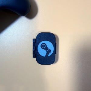 May include: A small, rectangular black plastic cover featuring a light blue Steam logo on the front surface. The cover has a hinged side mechanism and a textured finish, designed to fit over a port or button on a gaming device.