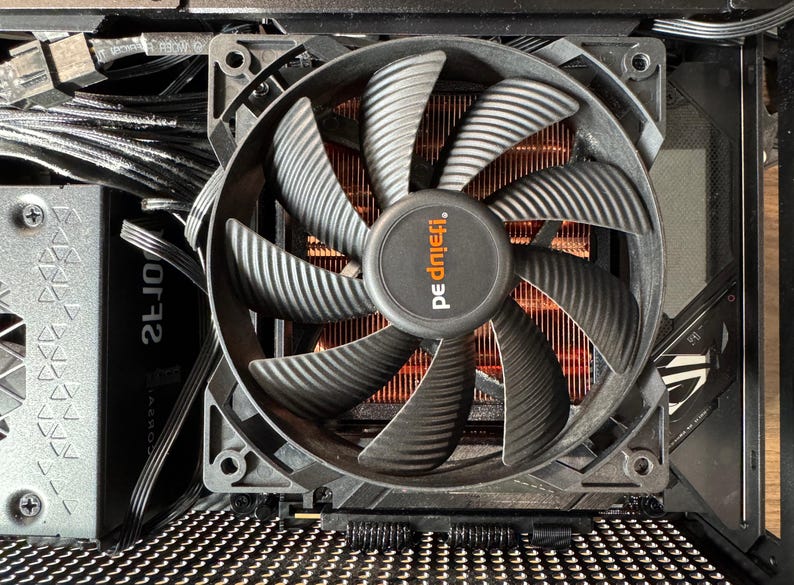 May include: A computer fan with black blades and a copper-colored heat sink. The fan has the brand name "be quiet!" printed on the center. The fan is installed inside a computer case with various wires and components visible.