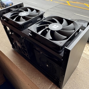 May include: A black computer case with two large cooling fans on top. The fans have black blades and a central logo. The case is rectangular and sits on a concrete ledge, with a street and parked cars in the background.
