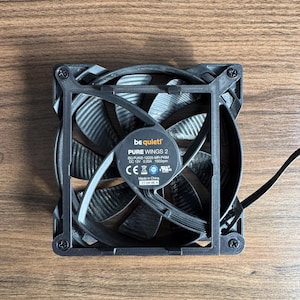 May include: A black computer fan with curved blades and a square frame. The fan has the text "be quiet! PURE WINGS 2" and other technical specifications printed on a label. The fan is on a wooden surface.