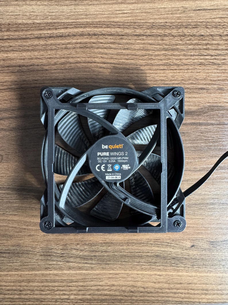 May include: A black computer fan with curved blades and a square frame. The fan has the text "be quiet! PURE WINGS 2" and other technical specifications printed on a label. The fan is on a wooden surface.
