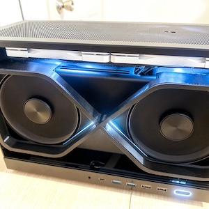 May include: A black computer case featuring a dual-fan cooling system with an X-shaped frame. The interior is illuminated by blue LED lighting, and the front panel includes multiple USB ports, a power button, and a headphone jack for connectivity.