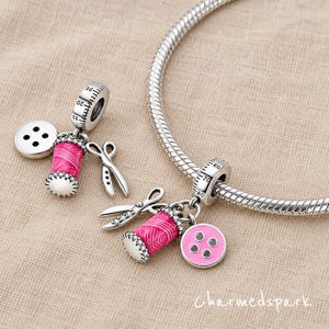 May include: Two silver charms featuring sewing themes, including a pink thread spool, a four-hole button, and scissors. One charm is attached to a silver snake chain bracelet. The metal components feature engraved ruler markings.