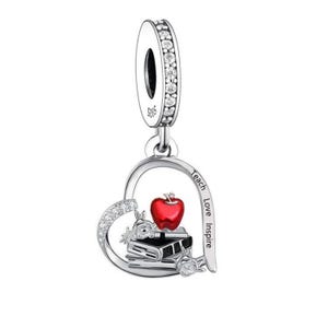 May include: A silver charm with a heart-shaped design. The charm features a red apple, books, and the words "Teach Love Inspire." The top loop is adorned with clear crystals, and the charm is designed for a bracelet or necklace.