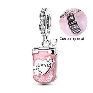 May include: A silver charm with a pink glittery design. The charm features a heart with the word "Love" and stars. The top of the charm has a ring with clear stones. A small charm of a flip phone is also shown. The text "Can be opened" is also shown.