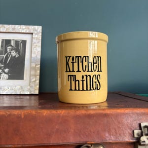 May include: A light yellow ceramic kitchen utensil holder with the words "KITCHEN THINGS" in black lettering. The holder is cylindrical. It is on a brown surface, with a framed black and white photo in the background.