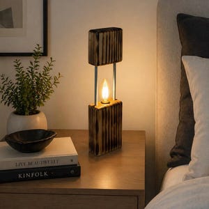 May include: A wooden table lamp with a warm, inviting glow. The lamp features a rectangular design with a light bulb in the center, supported by vertical metal rods. The lamp is placed on a wooden nightstand next to a bed.