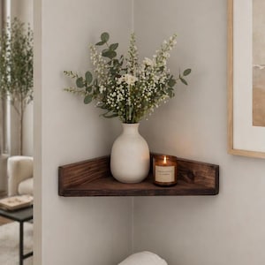 May include: A dark brown wooden corner shelf mounted on a wall. On the shelf sits a matte white ceramic vase filled with green eucalyptus stems and small white flowers, alongside a brown glass candle jar with a label reading Sandalwood & Amber.
