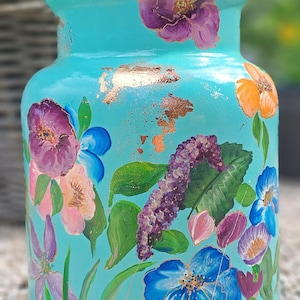 Op de afbeelding: Een handbeschilderde, turquoise keramische vaas versierd met kleurrijke bloemmotieven. De bloemen omvatten paarse, blauwe en oranje bloemen met groene bladeren. Goudblad accenten voegen een vleugje elegantie toe aan de decoratieve vaas.