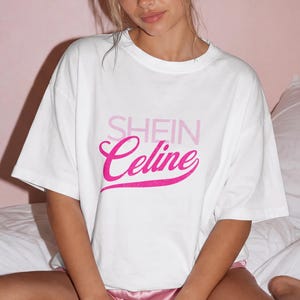May include: A white short-sleeved t-shirt featuring the text SHEIN in light pink block letters above the word Celine in a bold, cursive, dark pink font. The shirt has a crew neckline and a loose, relaxed fit, paired with pink satin shorts.