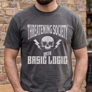 May include: Pepper-colored t-shirt with the text "Threatening Society with Basic Logic." The design features a skull wearing glasses and lightning bolts. The shirt is a short-sleeved crew neck.