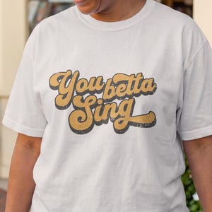 May include: A white short-sleeve t-shirt featuring the text "You betta Sing" in a bold, retro-style brown font with a distressed texture. The shirt has a crew neckline and a relaxed fit, providing a simple and clean look for casual wear.