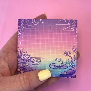 Op de afbeelding: Vierkante plaknotitie met een gradiëntachtergrond van paars, roze en geel. De illustratie toont een vijver met een kikker op een waterlelie, wolken, een maan en sterren. De notitie heeft een rasterpatroon.