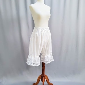 May include: White bloomers with a lace trim, displayed on a mannequin. The bloomers are made of a soft, lightweight fabric and have a ruffled lace detail at the hem. The mannequin is on a wooden stand against a light gray backdrop.