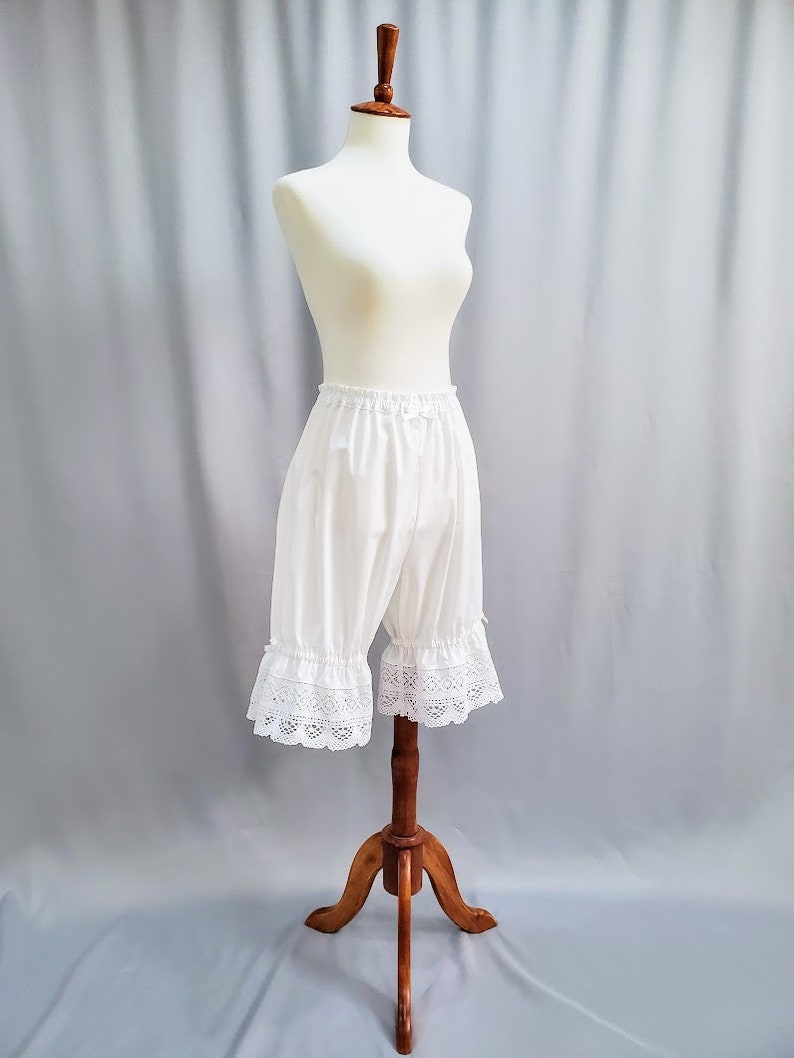 May include: White bloomers with a lace trim, displayed on a mannequin. The bloomers are made of a soft, lightweight fabric and have a ruffled lace detail at the hem. The mannequin is on a wooden stand against a light gray backdrop.