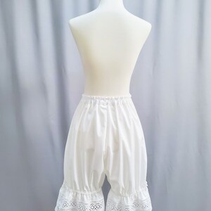 May include: White bloomers with a ruffled lace trim, displayed on a mannequin. The bloomers have a gathered waist and a loose, comfortable fit. The lace trim adds a delicate touch to the garment.