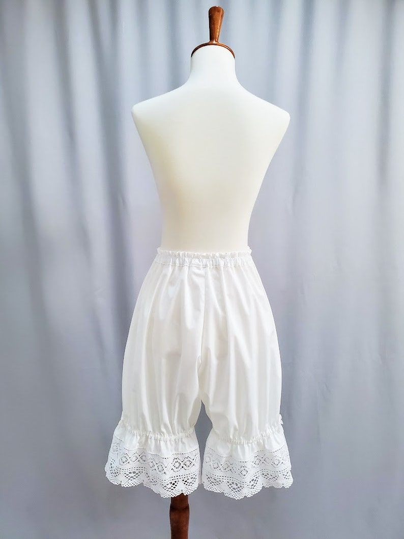 May include: White bloomers with a ruffled lace trim, displayed on a mannequin. The bloomers have a gathered waist and a loose, comfortable fit. The lace trim adds a delicate touch to the garment.