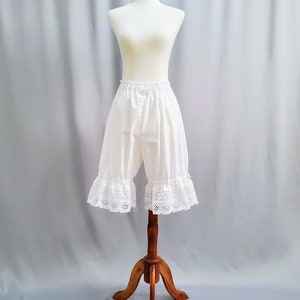 May include: White bloomers with a ruffled lace trim, displayed on a mannequin. The shorts have an elastic waistband and a loose fit. The garment is made of a lightweight fabric, perfect for historical reenactment or costume.