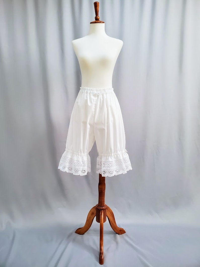 May include: White bloomers with a ruffled lace trim, displayed on a mannequin. The shorts have an elastic waistband and a loose fit. The garment is made of a lightweight fabric, perfect for historical reenactment or costume.