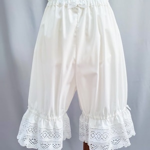 May include: White bloomers with an elastic waistband and a small bow detail. The bloomers feature a ruffled hem with intricate white lace trim. The fabric appears to be a soft, opaque material.