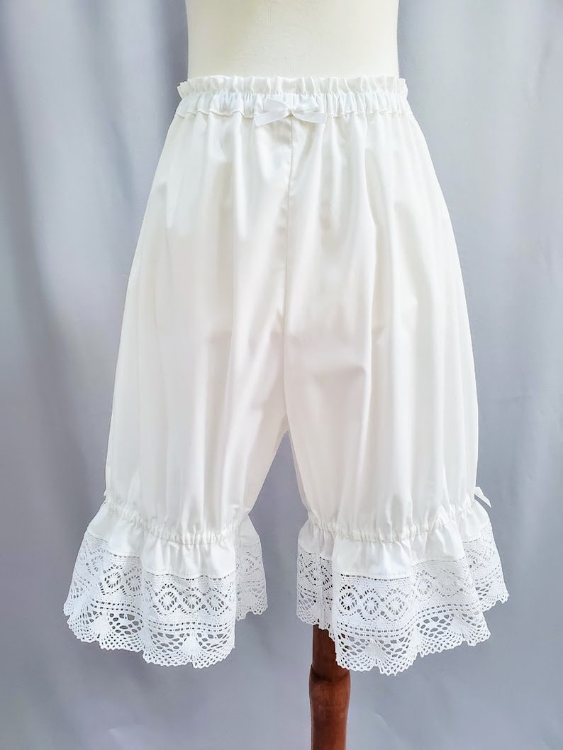 May include: White bloomers with an elastic waistband and a small bow detail. The bloomers feature a ruffled hem with intricate white lace trim. The fabric appears to be a soft, opaque material.