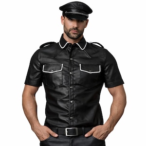 Pode incluir: Camisa preta de couro de manga curta com acabamento branco na gola e nos bolsos. A camisa tem uma frente abotoada e ombreiras. Uma boina de couro preta com corrente prateada tamb&eacute;m &eacute; vis&iacute;vel.