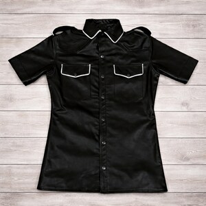Pode incluir: Camisa preta de manga curta com bot&otilde;es, dois bolsos no peito e gola. A camisa tem acabamento branco na gola e nos bolsos. A pe&ccedil;a &eacute; feita de material brilhante e tem ombreiras nos ombros.