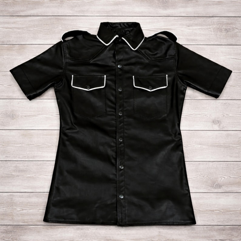 Pode incluir: Camisa preta de manga curta com bot&otilde;es, dois bolsos no peito e gola. A camisa tem acabamento branco na gola e nos bolsos. A pe&ccedil;a &eacute; feita de material brilhante e tem ombreiras nos ombros.