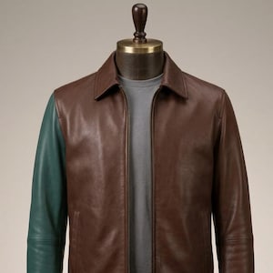 Peut inclure: Une veste en cuir marron avec une manche vert foncé contrastante, un col classique et une fermeture éclair frontale intégrale. La veste est présentée sur un mannequin portant un t-shirt gris à col rond sous le vêtement ouvert.