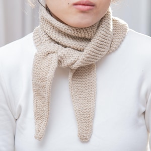 Può includere: Sciarpa beige lavorata a maglia di forma triangolare. La sciarpa è realizzata con un filato spesso e presenta una trama a coste. La sciarpa è avvolta attorno al collo e pende davanti.