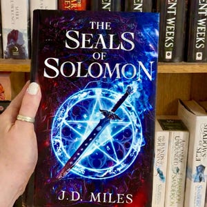 Może przedstawiać: Książka zatytułowana "The Seals of Solomon" z niebiesko-fioletową okładką przedstawiającą miecz i świecący pentagram. Widoczne jest nazwisko autora, J.D. Milesa. Obraz zawiera również tekst "PRE-ORDER SIGNED COPIES".