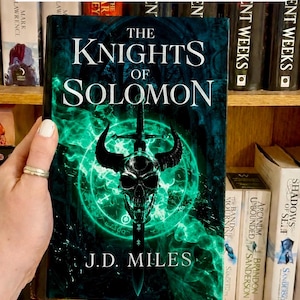 May include: A hardback book titled The Knights of Solomon by J.D. Miles with a dark cover featuring a glowing green skull and sword graphic. The text PRE-ORDER SIGNED COPIES appears in white boxes at the top and bottom of the image frame.