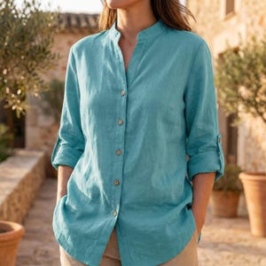 Può includere: Una camicia di lino color turchese con colletto alla coreana e abbottonatura frontale. Le maniche lunghe possono essere arrotolate e fissate con un cinturino abbottonato. La camicia è indossata con pantaloni beige.