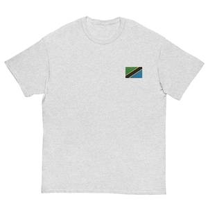 May include: A light gray t-shirt featuring the flag of Tanzania embroidered on the left chest. The flag design includes green, black, yellow, and blue colors. The shirt has a crew neck and short sleeves.
